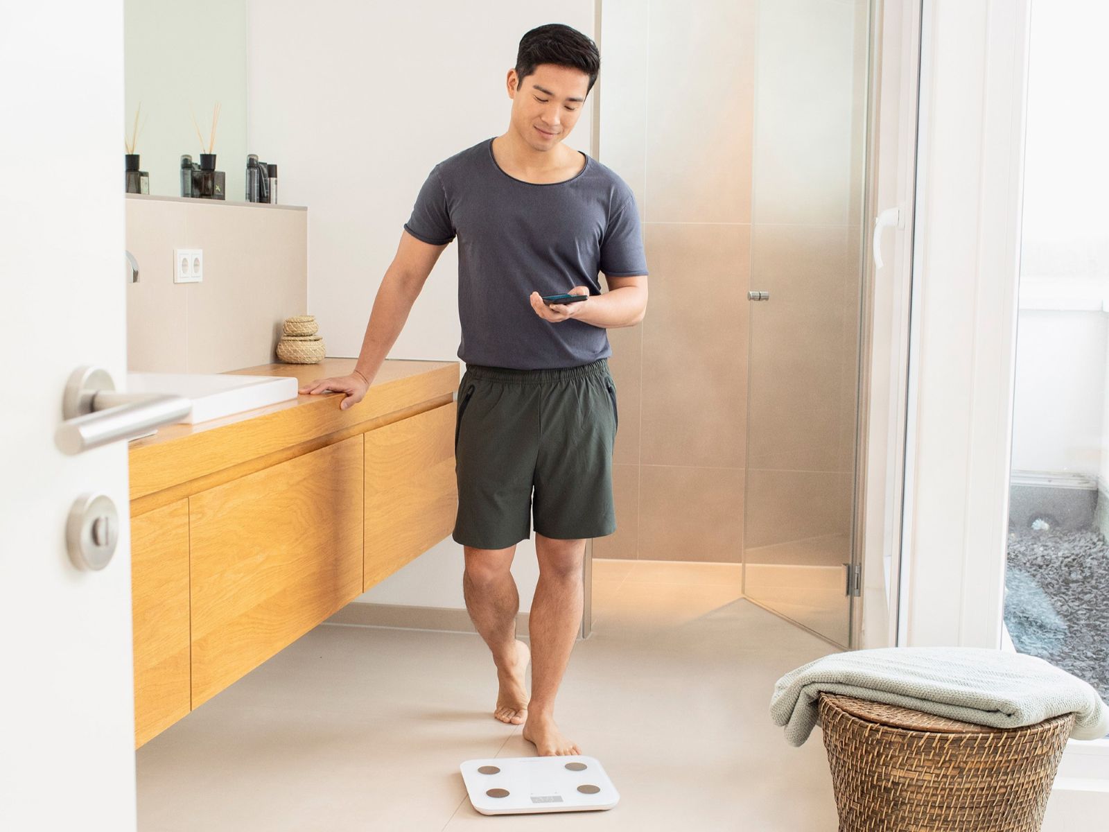 Ein Mann steht neben einer weißen Smart-Körperwaage im Badezimmer und schaut auf sein Smartphone, um seine Messdaten abzurufen.